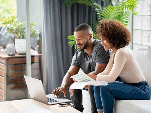 A couple sits on a white couch looking at a laptop while looking over document,