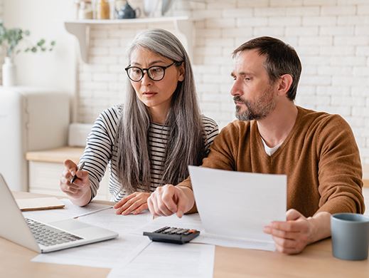 A woman and a man sit at a coffee table with a silver laptop, calculator and paperwork.