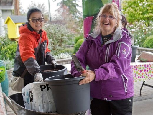 Two women with jackets on outside putting dirt from a wheelbarrow into two large pots